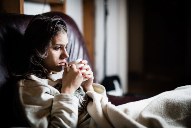 A young woman sits in bed, holding a warm drink, rugged up in blankets, with a red nose and a tissue.