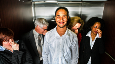 A smiling young man in a lift surrounded by four people holding their noses