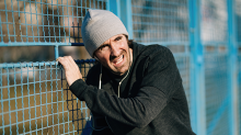 A breathless man in a black hoodie, wearing a grey beanie and white in-ear earphones holds onto a fence for support