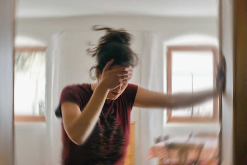 A woman stands into a doorway leaning heavily against the doorframe with her left hand and holding her forehead with her right hand. Her eyes are shut and the photo is swirly indicating dizzyness