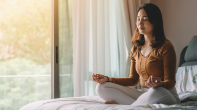 A woman sits on her bed in a meditation pose with her eyes closed and with upturned hands resting on her knees, thumbs and middle fingers touching
