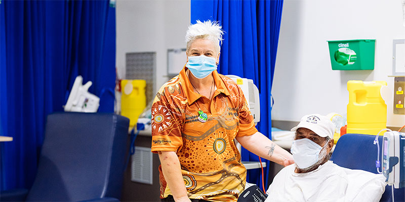 A woman in a face mask stands beside a man lying in a hospital bed, conveying a scene of care and concern.