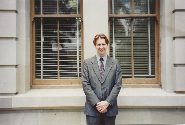 A young Dr Ross stands with his arms linked in front of him while he smiles at the camerain a suit.