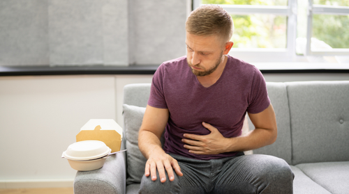 A man sits on a couch belching and holding his stomach. On the arm of the couch are fast-food containers.