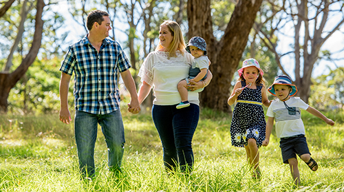 A young family with two children and a baby walk in a green field in front of some native trees