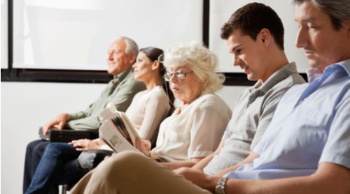 A line of people sit on chairs in a doctor's waiting room.