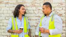 Two men in high visibility vests holding takeaway coffee cups stand facing each other in front of a wall chatting while on a coffee break