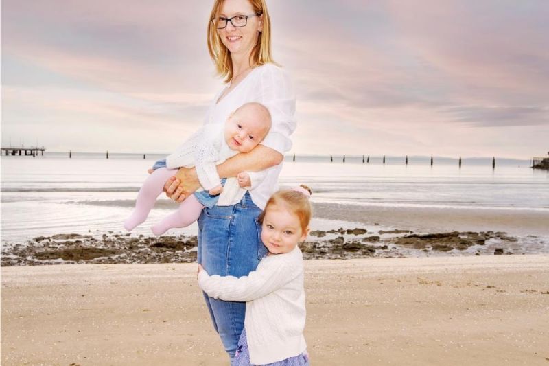 A mother on a beach with her two young children. She is holding the baby in her arms