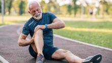 An athletic senior man dressed in running wear and shoes sits on an athletics track holding his right knee in both hands. He is grimacing in pain