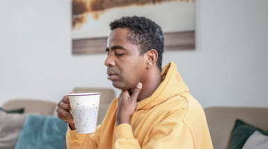 A man in a mustard-coloured hoodie sits on his couch with a cup of tea, holding his throat in pain due to acid reflux