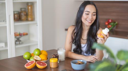 A young woman sits at her breakfast table holding and looking at a plastic bottle of multivitamin pills. In front of her on the table are a glass of milk, a jar of peanut butter, a bowl of wheat cereal, and a variety of fresh fruits