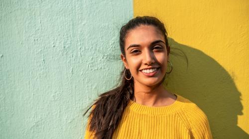 A smiling teenage girl in a mustard sweater standing in front of a roughly textured mustard and mint green wall. Her features are highlighted by a low sun from the right, casting a shadow to her left.