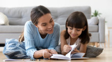 A mother and young daughter lying on the lounge floor, smiling and reading a book about sex education together